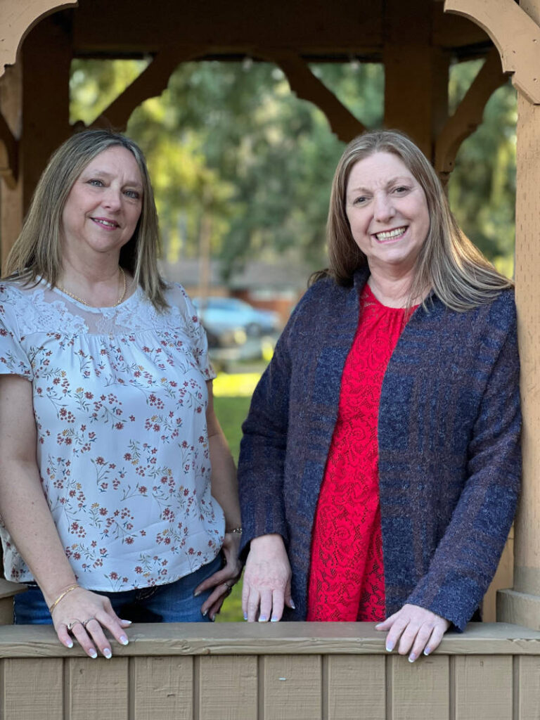 Two women standing in a gazebo. Laura, on left, shoulder length blond/brown hair, smiling. Julie, on the right, medium length blond/brown hair, with big smile.